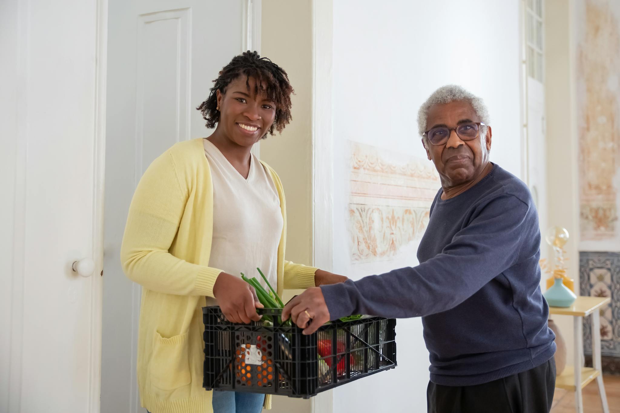 Smiling woman assists a senior man with food delivery indoors, promoting community support and care.